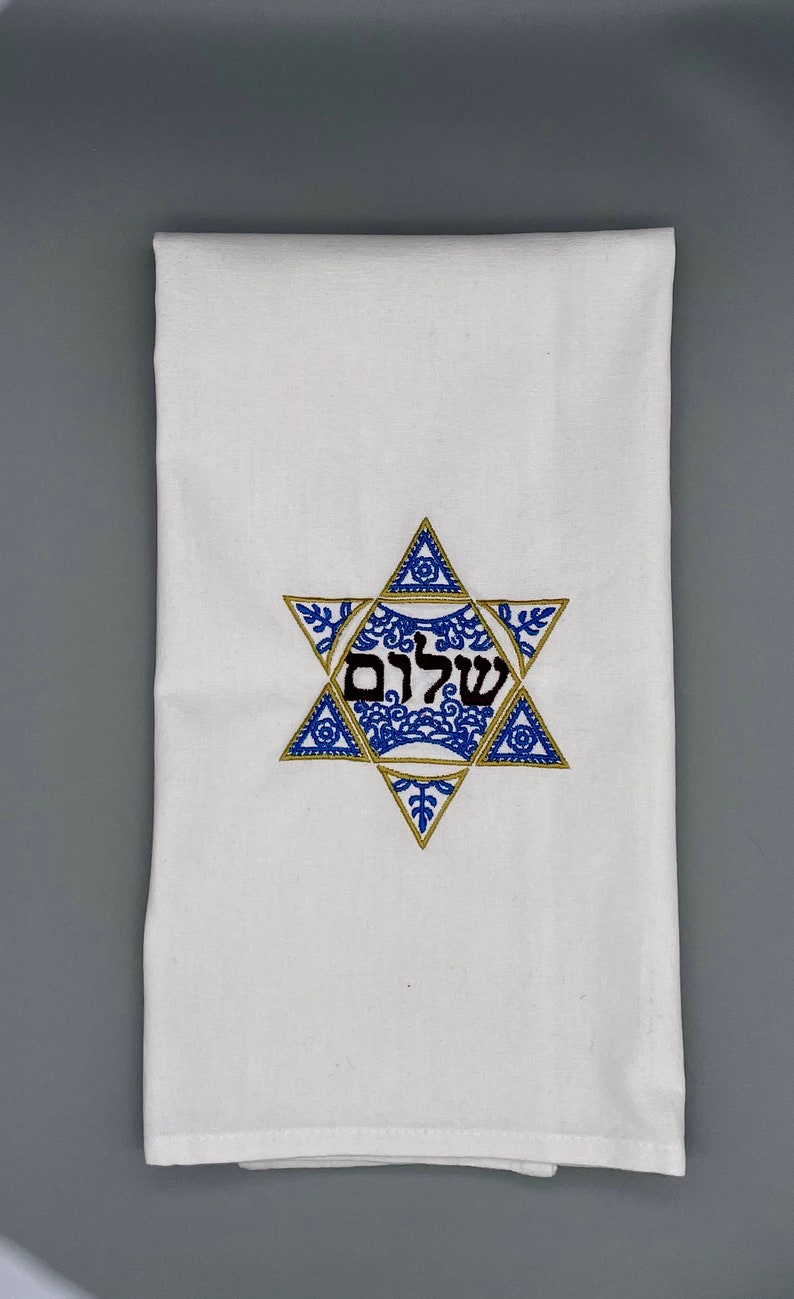 May include: White kitchen towel with a blue and gold embroidered Star of David with the Hebrew word "Shalom" in the center.