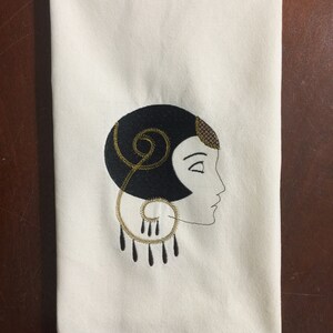 May include: A white kitchen towel with an embroidered design of a woman's face in profile. The woman has black hair and is wearing a gold earring. The design is in black and gold thread.