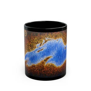May include: Black ceramic coffee mug featuring a vibrant abstract design. The artwork depicts a blue water body surrounded by brown and gold land masses. The mug has a black interior and rim, creating a striking contrast.