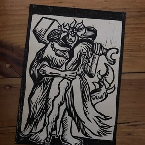 Op de afbeelding: Zwart-witte linosnede van een grote, gehoornde figuur die een kleinere figuur vasthoudt. De grotere figuur draagt een kroon en houdt een hamer vast. De kleinere figuur draagt een lange jurk.