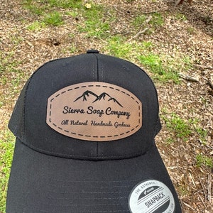 Peut inclure: Casquette de camionneur noire avec un patch en cuir marron qui dit "Sierra Soap Company All Natural Handmade Goodness" et un graphique de chaîne de montagnes. La casquette a un dos en maille noire et une étiquette argentée "The Authentic Yupoong Snapback".