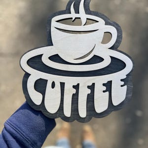 Puede incluir: Un letrero de madera con un diseño de taza de café y platillo. La palabra "COFFEE" está escrita en una fuente estilizada debajo de la taza.