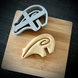 May include: A gray plastic cookie cutter shaped like a helmet and a beige cookie shaped like a helmet on a wooden cutting board.