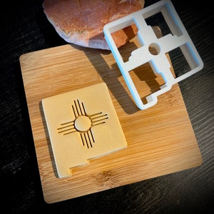 May include: A white plastic cookie cutter shaped like the state of New Mexico with the Zia symbol inside. The cookie cutter is on a wooden cutting board with a cookie that has been cut out using the cookie cutter.