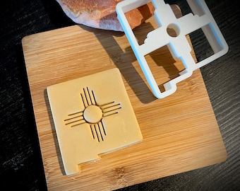 Könnte beinhalten: Ein weißer Plastik-Ausstecher in Form des Bundesstaates New Mexico mit dem Zia-Symbol im Inneren. Der Ausstecher liegt auf einem Holzbrett mit einem Keks, der mit dem Ausstecher ausgestochen wurde.
