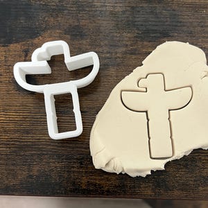 May include: A white plastic cookie cutter in the shape of a totem pole is next to a piece of beige dough with a totem pole imprint. The cookie cutter has a rectangular base and a detailed top with curved lines.