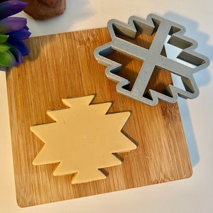 May include: Two cookie cutters on a wooden cutting board. The cookie cutters are both star-shaped with a geometric design. One cookie cutter is silver and the other is beige.