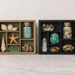 May include: Two square wooden shadow boxes, one tan and one black, each with six compartments. The tan box holds seashells, a starfish, and a jar of blue sea glass. The black box displays crystals and a jar of colorful stones.