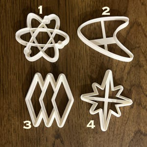 May include: Four white plastic cookie cutters, numbered 1 through 4. The cutters are shaped like an atom, a spaceship, a set of three diamonds, and a star.