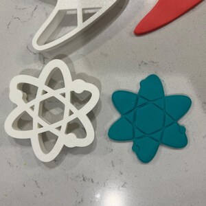 以下が含まれることがあります： 原子形の白いプラスチック製のクッキー型が2つ。1つはターコイズブルーの原子が入っています。