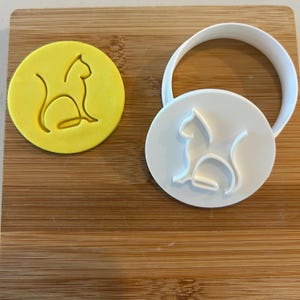 May include: A white plastic cookie cutter in the shape of a cat sitting down. The cutter is on a wooden surface next to a yellow cookie dough cutout of the same cat shape.
