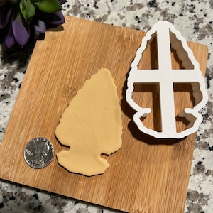 May include: A white plastic cookie cutter shaped like an arrowhead, with a brown cookie dough cutout next to it on a wooden cutting board.