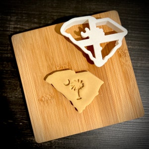 May include: A white plastic cookie cutter in the shape of the state of South Carolina. A brown cookie in the shape of South Carolina with a crescent moon and palmetto tree design is on a wooden cutting board.