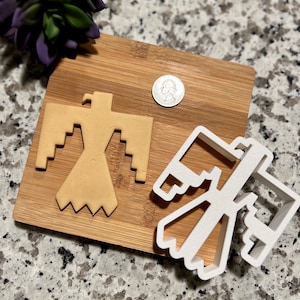 May include: A white plastic cookie cutter in the shape of a stylized Native American eagle design. A cookie cut out using the cutter is shown on a wooden cutting board.