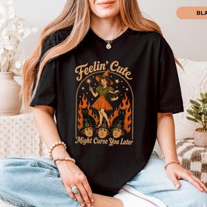 Puede incluir: Una camiseta negra de manga corta con un estampado gráfico de una bruja sosteniendo una varita y un libro, rodeada de llamas, dos gatos negros y tres rostros con sombreros de peregrino. El texto dice Feelin' Cute Might Curse You Later.