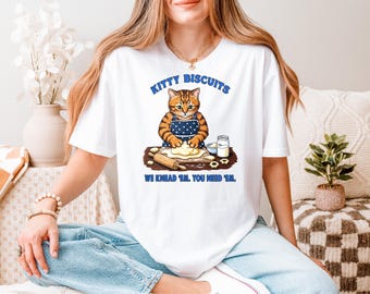 May include: White t-shirt featuring a cartoon cat wearing an apron and kneading dough. The text "KITTY BISCUITS" is above the cat, with "WE KNEAD 'EM. YOU NEED 'EM." below. The cat is orange and brown, and the apron is blue with white dots.