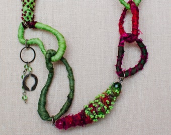May include: A handmade necklace with green and pink fabric, beads, and metal accents. The necklace features a unique design with loops and knots.