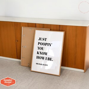 Op de afbeelding: Een witte ingelijste print met de tekst "Just poopin' you know how I be." en "Michael Scott" onderaan. De print leunt tegen een houten kast.