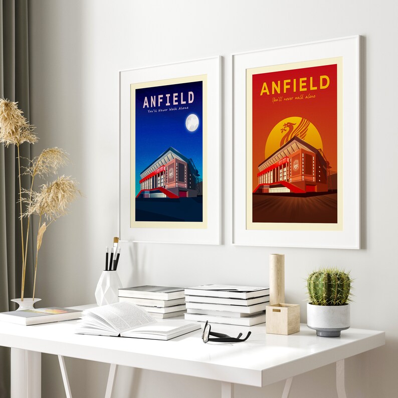 May include: Two framed art prints featuring Anfield stadium. One depicts a night scene with a full moon and the text "ANFIELD" and "You'll Never Walk Alone." The other shows a sunset with a Liver bird and the same text. Both are in white frames.