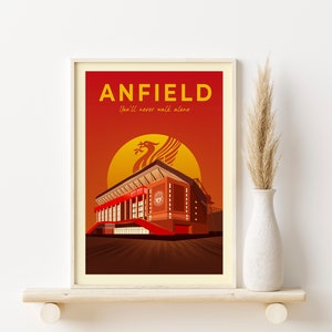 May include: Framed art print featuring a stylized illustration of Anfield stadium. The artwork has a red and orange gradient background with the word "ANFIELD" in yellow. The Liverpool FC emblem is visible.