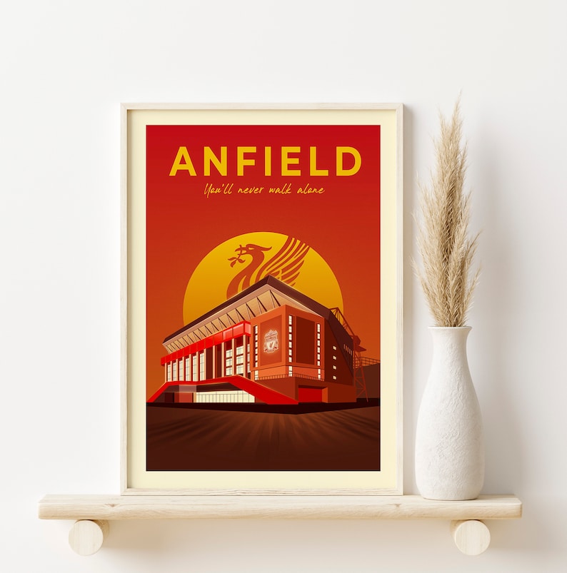May include: Framed art print featuring a stylized illustration of Anfield stadium. The artwork has a red and orange gradient background with the word "ANFIELD" in yellow. The Liverpool FC emblem is visible.