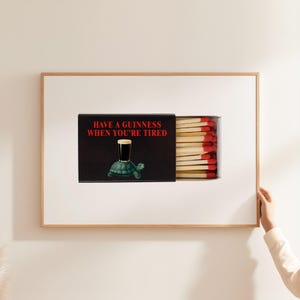 May include: A framed print featuring a matchbox design. The matchbox is black with the text "HAVE A GUINNESS WHEN YOU'RE TIRED" in red. The matchbox illustration includes a pint of Guinness on a turtle. The matches have red tips.