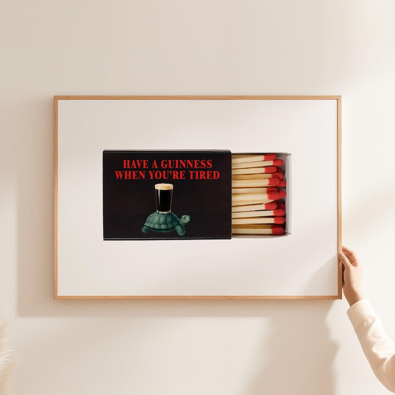 May include: A framed print featuring a matchbox design. The matchbox is black with the text "HAVE A GUINNESS WHEN YOU'RE TIRED" in red. The matchbox illustration includes a pint of Guinness on a turtle. The matches have red tips.