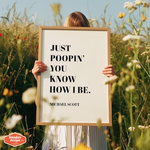 Op de afbeelding: Een witte geframed poster met zwarte tekst die "JUST POOPIN' YOU KNOW HOW I BE. -MICHAEL SCOTT" luidt. De poster wordt vastgehouden door een persoon in een veld met bloemen.