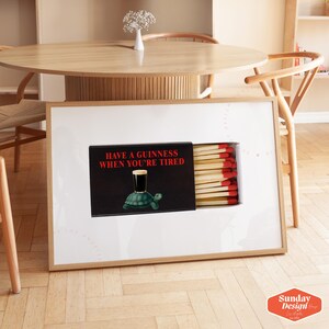 May include: A framed art print featuring a matchbox design with the text "HAVE A GUINNESS WHEN YOU'RE TIRED." The artwork includes a Guinness glass on a turtle. The matchsticks have red tips. The frame is light wood.