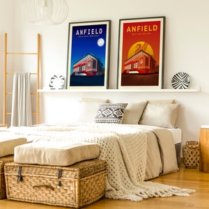 May include: Two framed Anfield stadium art prints. One features a night scene with a full moon, the other a sunset. Both have the text "ANFIELD" and "You'll Never Walk Alone". The prints are displayed above a bed.