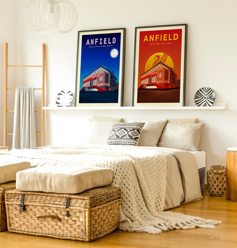 May include: Two framed Anfield stadium art prints. One features a night scene with a full moon, the other a sunset. Both have the text "ANFIELD" and "You'll Never Walk Alone". The prints are displayed above a bed.