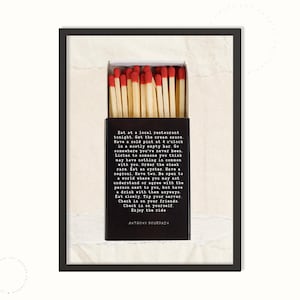 May include: A framed print featuring a box of matches with red tips, set against a textured, off-white background. The matchbox displays a quote by Anthony Bourdain in a black font. The frame is black.