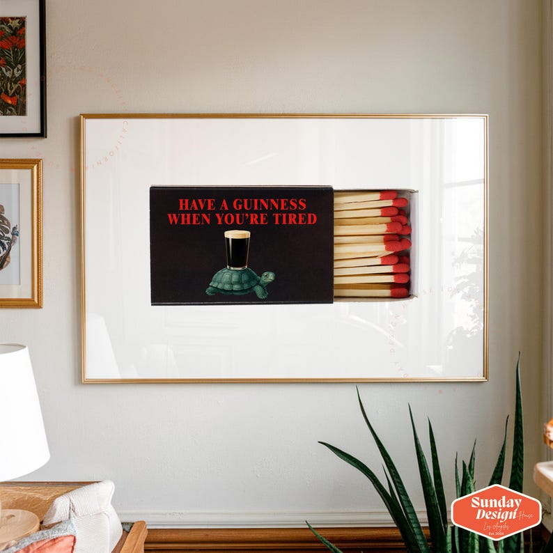 May include: Framed artwork featuring a matchbox design. The matchbox has the text "HAVE A GUINNESS WHEN YOU'RE TIRED" in red. The image on the matchbox shows a pint of Guinness on a turtle. The matches are visible.