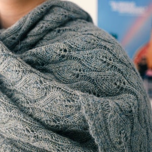 Könnte beinhalten: Ein weicher, grauer Strick Schal mit einem strukturierten Muster. Der Schal ist gefaltet und über die Schulter einer Person gelegt.