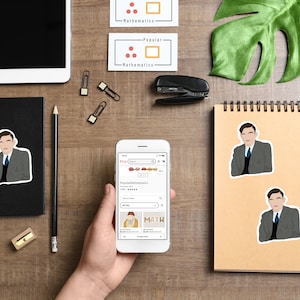 Puede incluir: Una persona sostiene un teléfono inteligente que muestra una pantalla con la aplicación Etsy abierta. La pantalla muestra una búsqueda de "Popular Mathematics" y dos listados para "MATH" y "Easy Mathematics".