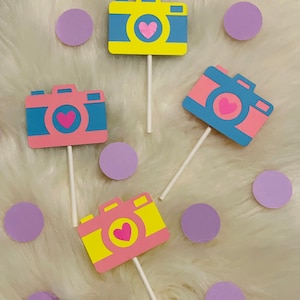 May include: Four paper camera cupcake toppers with pink and yellow bodies and blue lens. Each camera has a pink heart in the center. The toppers are on white sticks and are surrounded by purple circles.