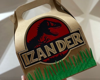May include: A brown cardboard favor box with a green grass border. The box features a red and black Jurassic Park logo with a dinosaur silhouette and the name "IZANDER" in gold lettering.