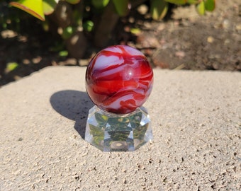 May include: A red and white swirled glass marble on a clear glass stand. The marble has a white swirl pattern on a red background.