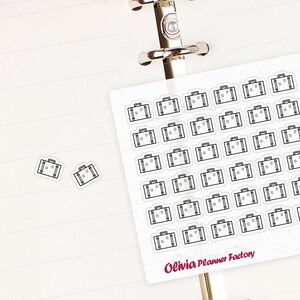May include: A sheet of white stickers with black outlines of suitcases. The stickers are arranged in a grid pattern. The sheet is labeled "Olivia Planner Factory" and "L329".