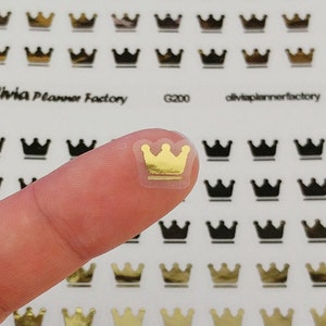 May include: A sheet of gold crown stickers. The stickers are on a white background and are arranged in rows. A finger is holding one of the stickers up to show the detail. The sticker is transparent with a gold crown inside.