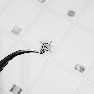 May include: A clear sticker with a black outline of a light bulb. The sticker is being held by tweezers.