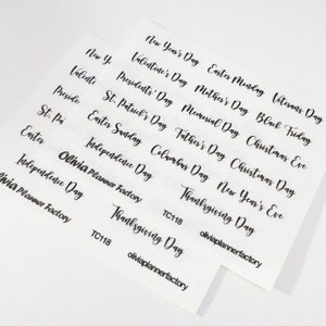 May include: A set of white stickers with black text listing various holidays, including New Year's Day, Valentine's Day, President's Day, St. Patrick's Day, Easter Sunday, Mother's Day, Memorial Day, Father's Day, Columbus Day, Thanksgiving Day, Christmas Day, Christmas Eve, New Year's Eve, Veterans Day, Black Friday, and Independence Day. The stickers also include the text "Olivia Planner Factory" and "TC118".