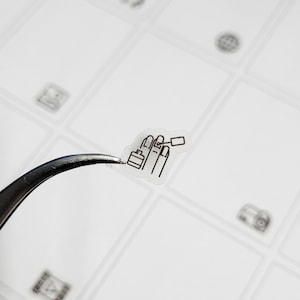 May include: A small, transparent sticker featuring a black line drawing of a hand with painted nails and nail polish. The sticker is held by a pair of silver tweezers against a white background with other small, faint icons.