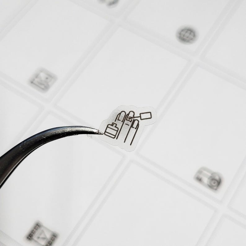 May include: A small, transparent sticker featuring a black line drawing of a hand with painted nails and nail polish. The sticker is held by a pair of silver tweezers against a white background with other small, faint icons.