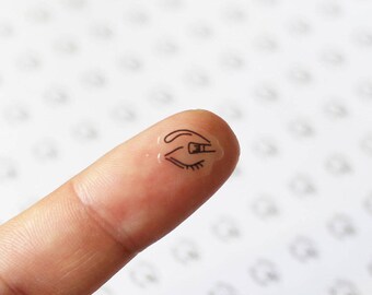 May include: A close-up of a fingertip with a clear sticker of an eye with an eyebrow and an eyeliner brush applying makeup.