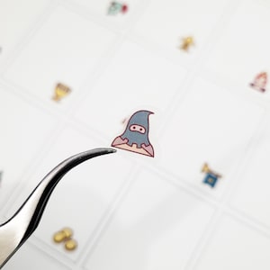 May include: A small, transparent sticker featuring a cartoon character in a blue hooded outfit with a red trim and a mask. The sticker is held by a pair of tweezers against a white background with other small stickers.