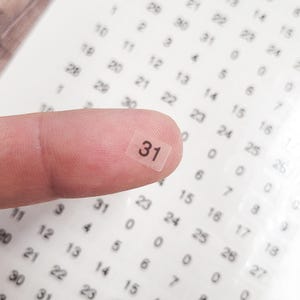May include: A finger holding a small, transparent square sticker with the number "31" printed in black. The sticker is surrounded by other numbered stickers on a white background. The image is a close-up.
