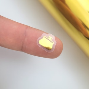 May include: A small, gold-colored purse sticker with a clear outline. The sticker is on a finger, with a gold roll in the background. The purse design is simple and elegant, suitable for scrapbooking or crafts.