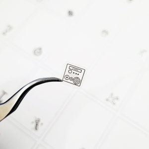 May include: A small, transparent sticker featuring a black and white line drawing of a stereo system. The sticker is held by silver tweezers against a white background with faint, scattered markings.