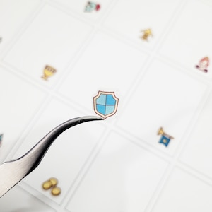 May include: A small, transparent sticker of a shield with a blue and brown design is held by tweezers. The shield has a blue cross on a light blue background. Other small stickers with various designs are visible in the background.